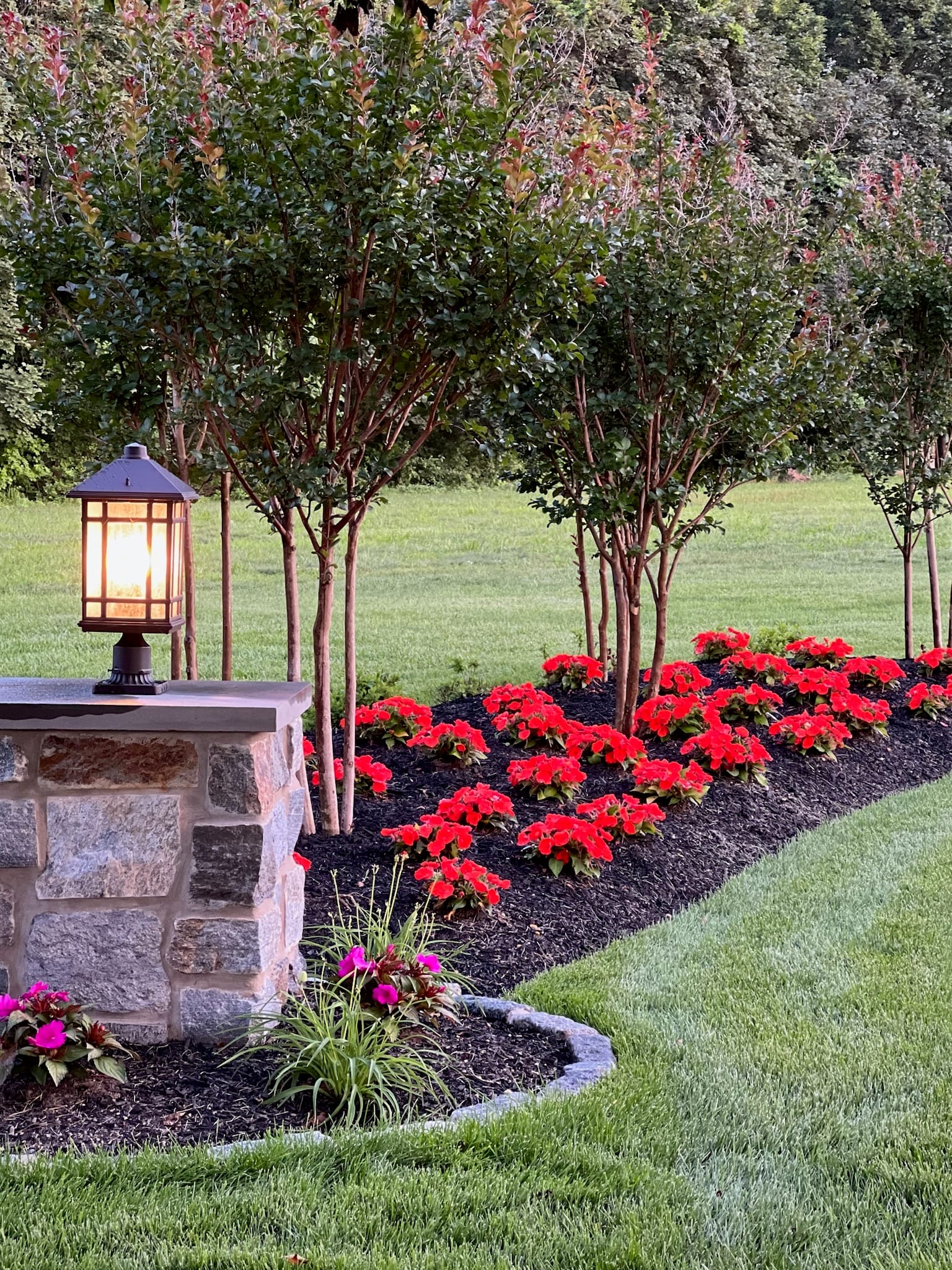 Stone pillar with lit craftsman lantern beside a bed of red flowers and crape myrtles