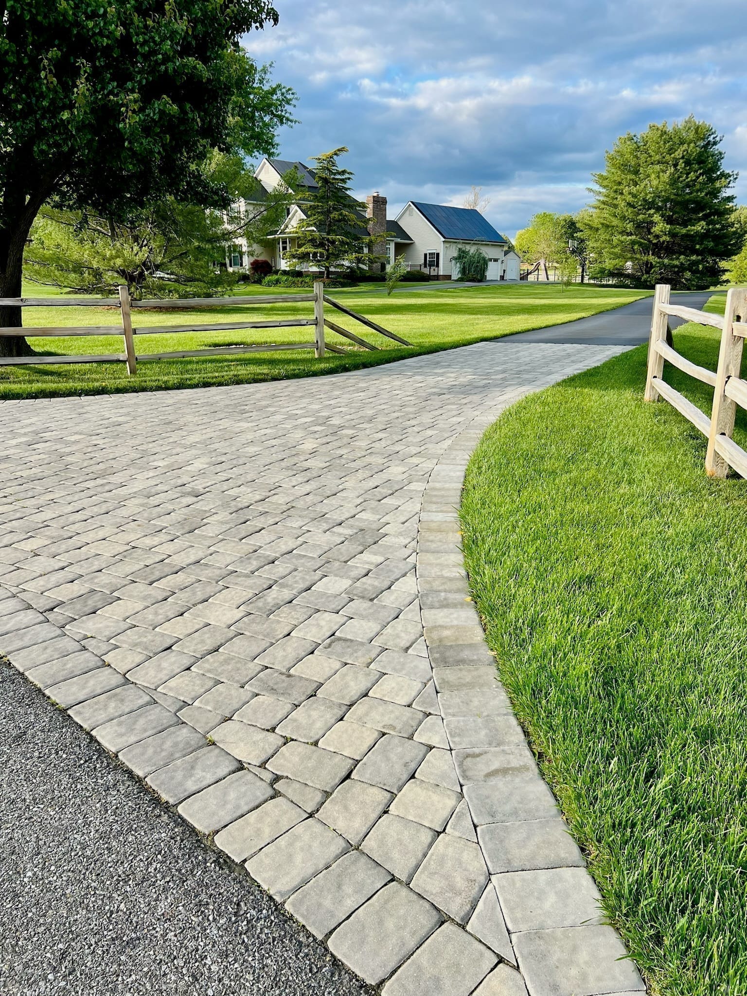 Curving Belgard paver driveway viewed from the side, bordered by lawn and split-rail fencing
