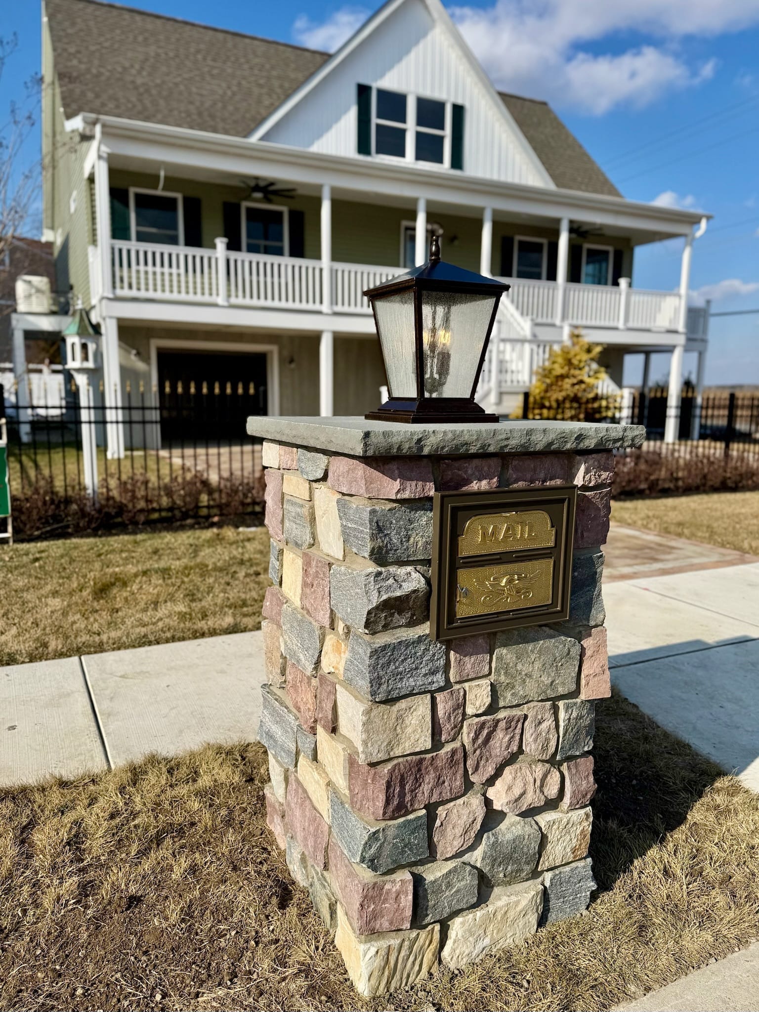 Stacked multi-color stone pillar with a brass mailbox door and craftsman lantern in front of a farmhouse-style home